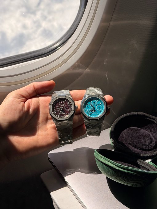 A hand holds two luxury watches—one with a dark dial and one with a bright blue dial—both wrapped in protective plastic, against an airplane window backdrop.