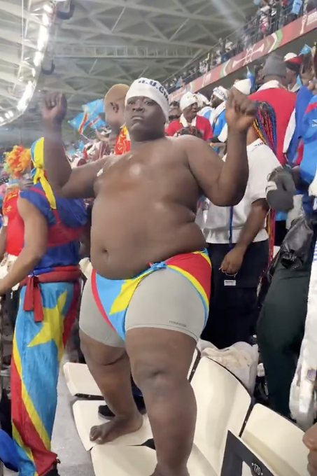The image features a man standing in front of a crowd at an event, wearing colorful shorts and a headband. He appears to be dancing or performing some sort of act for the audience. The crowd consists of people from various backgrounds, all gathered together to enjoy the show.