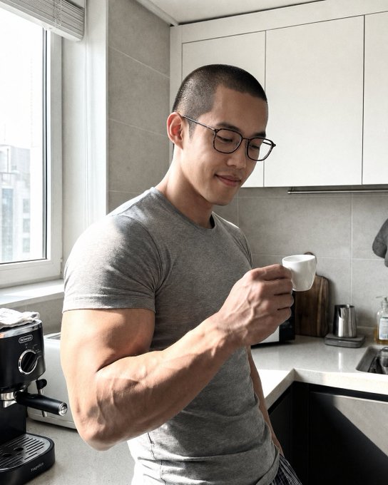 A muscular man wearing glasses and a gray t-shirt is smiling while holding a small white espresso cup in a modern kitchen with an espresso machine on the counter.