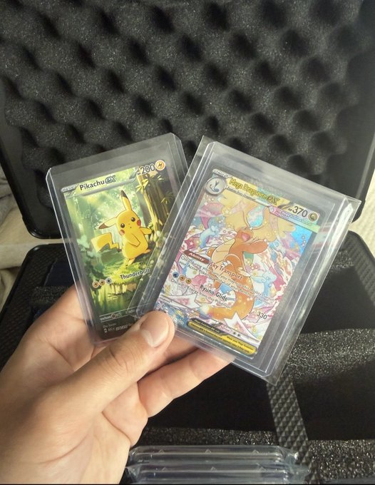 Hand holding two graded Pokémon cards in protective slabs: a Pikachu EX card and a Mega Blaziken ex card, displayed in front of a foam-lined case.