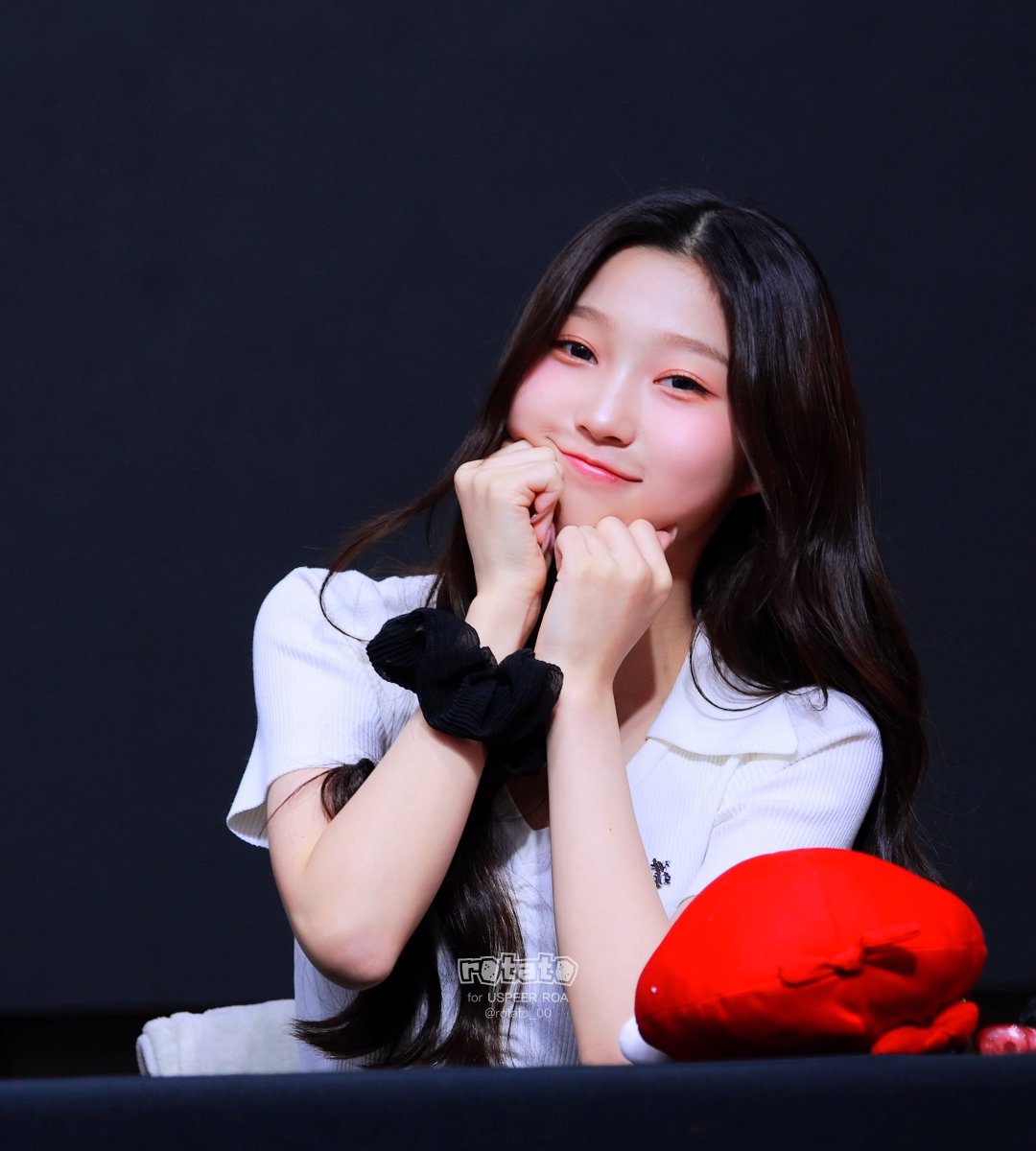 A smiling young woman with long dark hair in a white top poses with her hands near her face, wearing a black scrunchie, with a red plush toy visible beside her.
