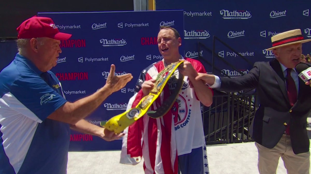 The image shows Joey Chestnut receiving the iconic Mustard Belt championship trophy at the Nathan's Hot Dog Eating Contest, flanked by the reigning champion in the red cap and another man in a straw boater hat, with sponsor logos for "Nathan's," "Polymarket," and "Chinet" visible on the backdrop, celebrating his record-breaking 18th win where he consumed 66 hot dogs in 10 minutes.