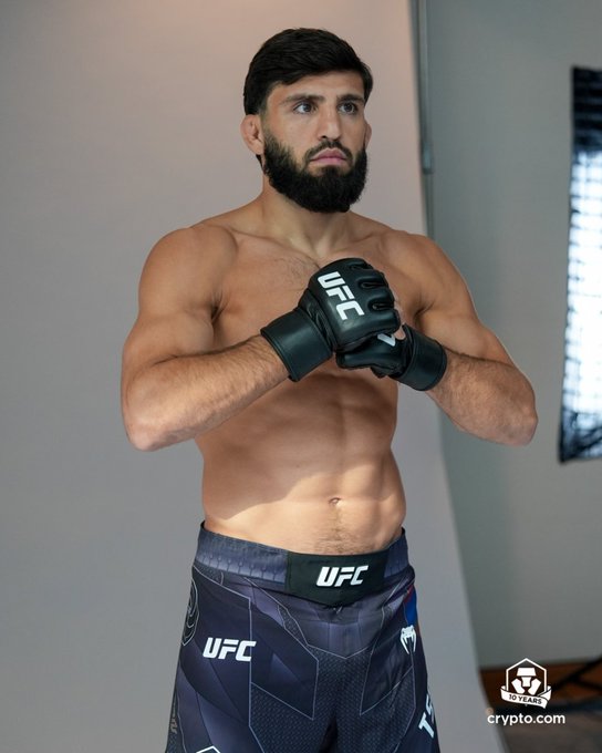 A shirtless UFC fighter with a beard poses in fighting stance wearing black UFC gloves and dark blue UFC-branded shorts, with a "Crypto.com 10 Years" watermark visible in the corner, promoting the UFC 331 media day event.