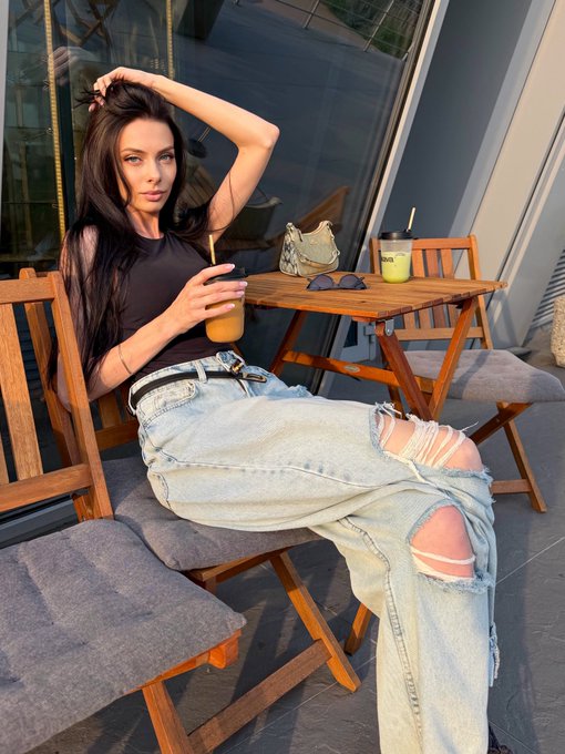 A woman with long dark hair sits at an outdoor cafe table wearing a black top and ripped light blue jeans, holding an iced coffee.