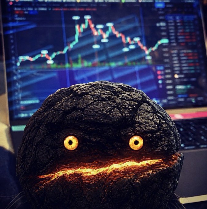 A glowing-eyed volcanic rock creature figurine positioned in front of a computer monitor displaying a cryptocurrency or stock trading chart with candlestick patterns.