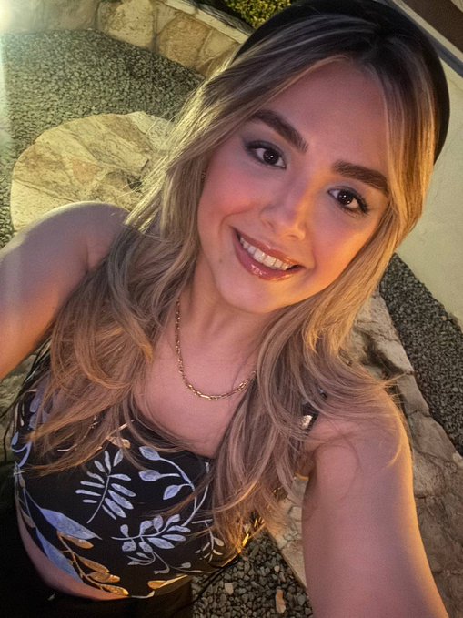 A selfie of a smiling young woman with long blonde hair, wearing a black top with leaf patterns and a gold necklace.