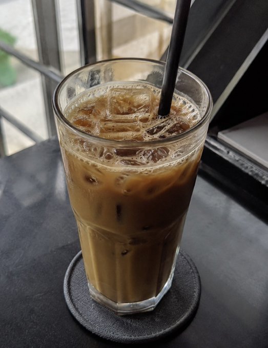 A glass of iced coffee with a black straw sitting on a coaster on a dark table near a window.