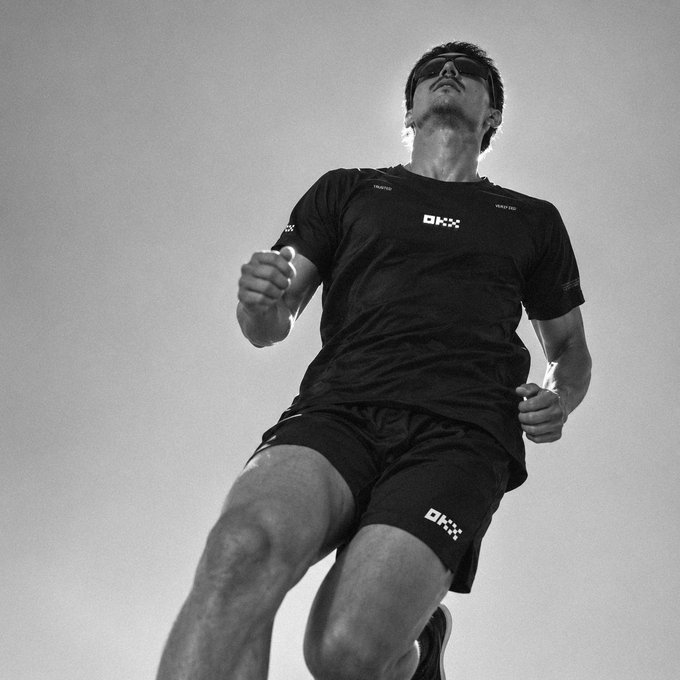 A black and white low-angle shot of an athlete mid-stride wearing sunglasses, a black t-shirt, and black shorts branded with the 'OK' logo, photographed against a plain light background.