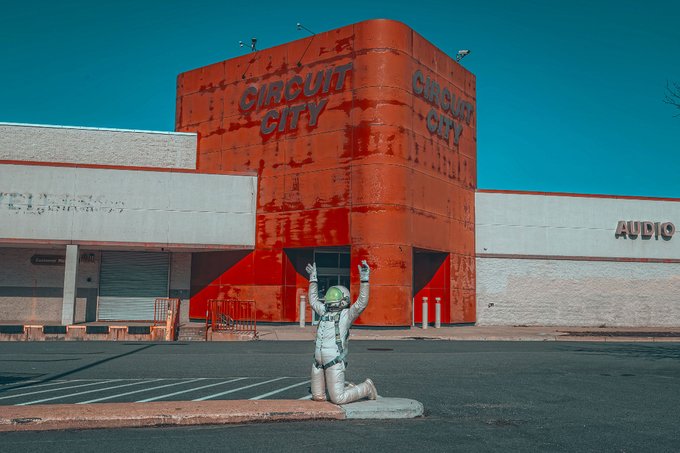 A photo of a weathered orange 'CIRCUIT CITY' storefront with an astronaut figure kneeling in the empty parking lot with arms raised.