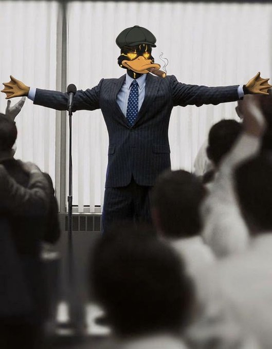 A digitally altered image of a corporate figure in a pinstripe suit with Daffy Duck's head wearing a flat cap, smoking a cigar and gesturing with outstretched arms before an audience.