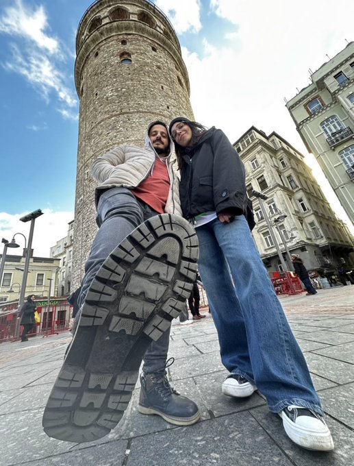 Two people posing in front of a tall stone tower (likely Galata Tower) with one person lifting their boot toward the camera in a low-angle shot.