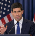 A man in a suit speaks at a podium, gesturing with his hand, with an American flag visible in the background.