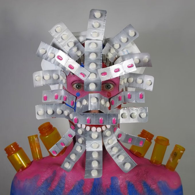 An artistic photograph showing a person whose face is obscured by an arrangement of blister packs of pills, with orange pill bottles displayed on their pink-painted shoulders.