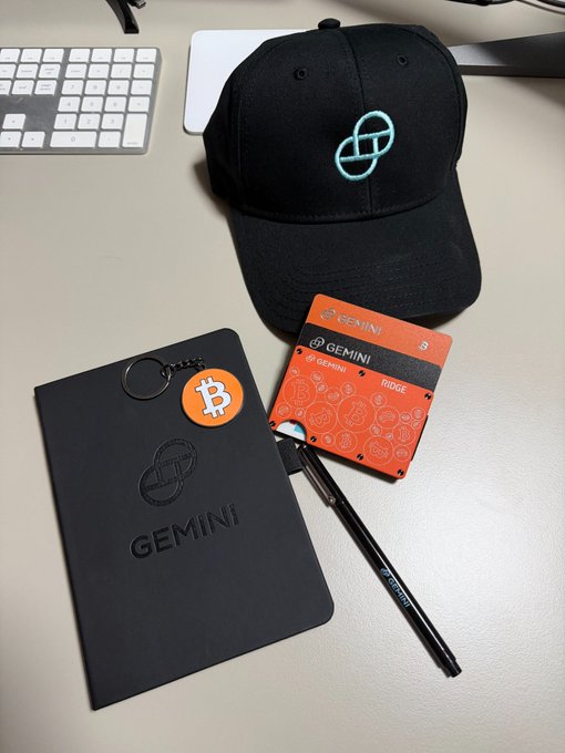 Gemini-branded merchandise is arranged on a desk, including a black cap with the cyan Gemini logo, a black Gemini folder/keychain with an orange Bitcoin charm, and two orange Gemini cards alongside a pen.