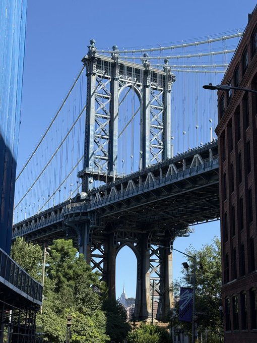 A street-level view of the Manhattan Bridge on a clear day, framed by modern and brick buildings.