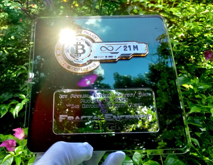 A commemorative crypto collectible titled "1st Annual Bitcoin Infinity Day, August 21, 2023, The Bitcoin Infinity Key by Fractal EnergyPU 8/21" featuring a Bitcoin branded key design, held outdoors against green foliage.