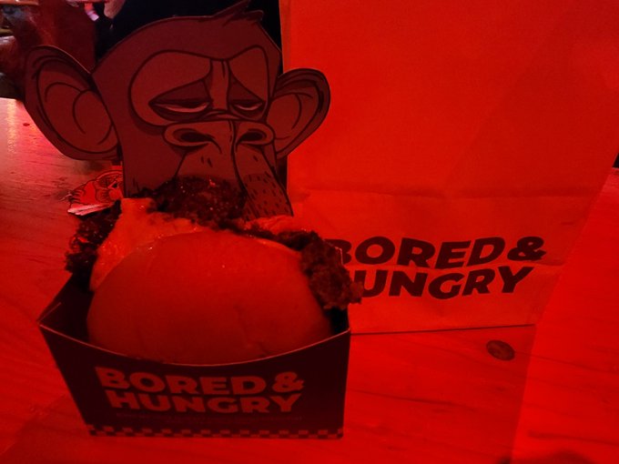 A hamburger in a 'BORED & HUNGRY' branded box under red lighting, with a cardboard cutout of an ape-like face standing behind it.
