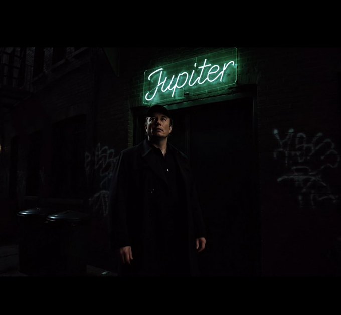 The image features a man standing outside, wearing a black coat and hat, with his back turned towards the camera. The background includes a neon sign that reads "Jupiter" and a building with graffiti on its facade. There are also trash cans visible in the scene. The image is related to an advertisement for the SpaceX IPO, which is set to launch tomorrow at $SPCX.