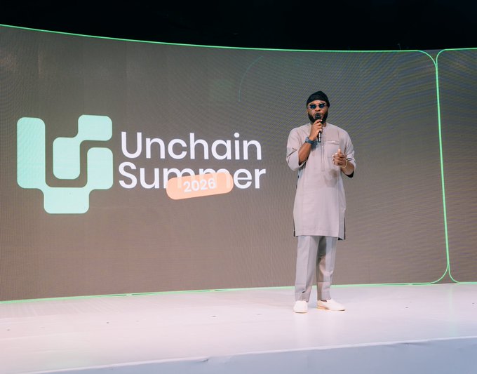 A speaker in a white traditional outfit addresses the audience at the "Unchain Summer 2026" event in Lagos, standing on stage in front of a large screen displaying the event's logo and branding. The moment captures the speaker's presentation during what appears to be a community-focused blockchain or tech gathering in Nigeria.