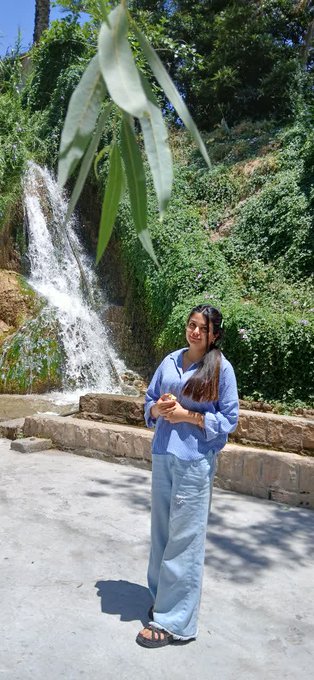 A woman in a blue shirt and jeans stands smiling near a cascading waterfall surrounded by lush greenery, capturing a serene morning moment that perfectly fits the cheerful "Gm" greeting.