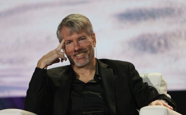 Michael Saylor, founder of Strategy (formerly MicroStrategy), smiles and gestures while seated during what appears to be a conference or speaking engagement, likely discussing the company's recent decision to sell 1,690 BTC ($186 million) at an average price of $64,262, reducing its holdings to 847,477 BTC to manage preferred stock structure and liquidity.