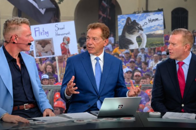 Three college football broadcasters sit at a desk with a green screen showing a crowd behind them, where fans have creatively inserted cat meme signs reading "Flirting" with "Sexual Harassment," and a dog pondering "Hmm..." with a thinking cat captioned "Hmm..." — directly referencing the $HMM meme coin promotion tied to getting a "Thinking Cat" sign on College Gameday.