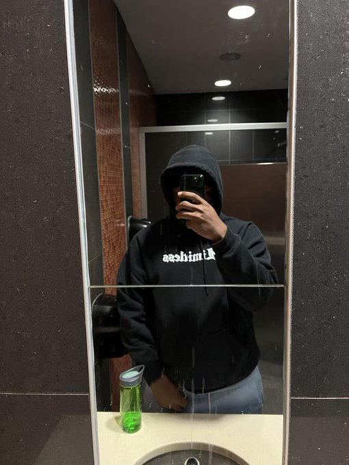A mirror selfie of a person wearing a black hoodie with mirrored text, holding a phone to take the photo, with a green water bottle on the counter.