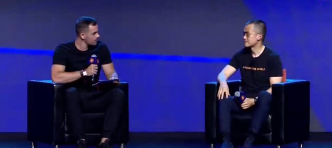 Two men seated in black chairs on a blue-lit stage during what appears to be an interview or talk show panel discussion.