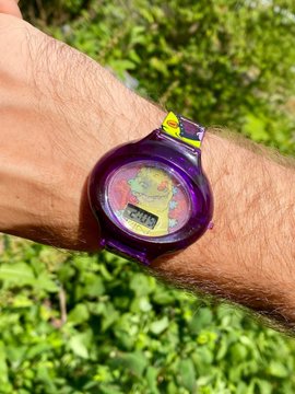 A close-up of a purple transparent digital wristwatch on someone's arm, with green vegetation in the background.