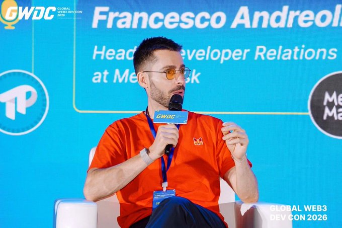 Francesco Andreoli, Head of Developer Relations at MetaMask, speaking at the Global Web3 Dev Con 2026 (GWDC). He is wearing an orange t-shirt, glasses, and holding a GWDC microphone.