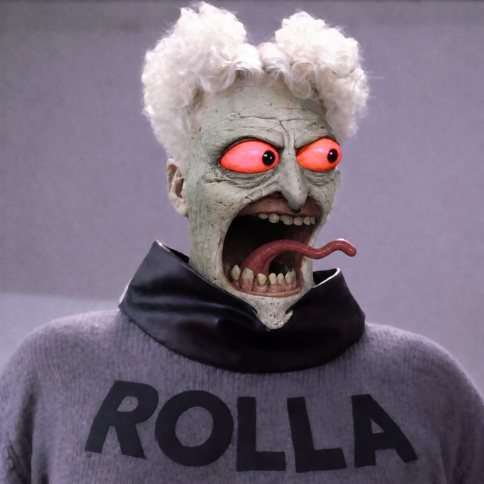 A bizarre mask with wild white hair, glowing orange eyes, and a protruding red tongue wears a gray sweater reading 'ROLLA'.
