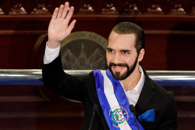 The image features a man wearing a blue and white striped shirt with his hand raised high in the air while waving. The background of the photo shows a wooden paneled room, which could be an auditorium or a similar setting for an event. The main focus is on the man's gesture and attire, suggesting that he might be attending a formal occasion or speaking at a public gathering related to Bitcoin education.
