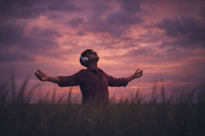 A person wearing headphones stands in a grassy field with arms outstretched, face tilted upward toward a dramatic pink and purple sunset sky.