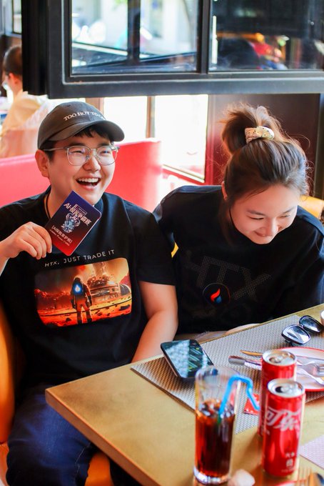 A man wearing a 'Tom & Jerry' cap and glasses, alongside a woman with her hair in a bun, smiling together at a restaurant table with Coca-Cola cans and a smartphone visible.