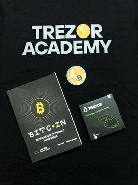A black t-shirt displays 'TREZOR ACADEMY' in white and yellow lettering, photographed with a physical Bitcoin token and two informational booklets, including one titled 'Bitcoin Definition of Money'.