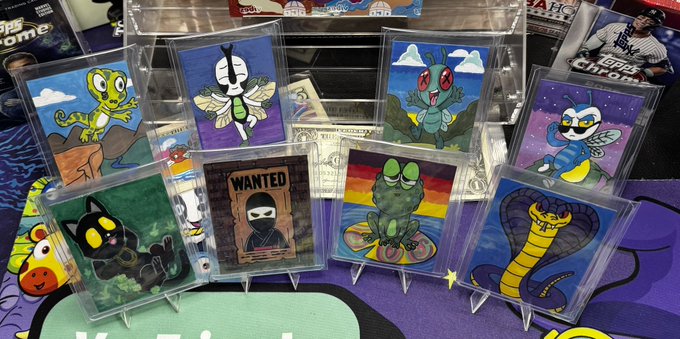 A display of handmade cartoon trading cards featuring insect-themed characters like a grasshopper, dragonfly, beetle, mosquito, snake, and cat, plus a 'WANTED' ninja poster, arranged on a colorful playmat.