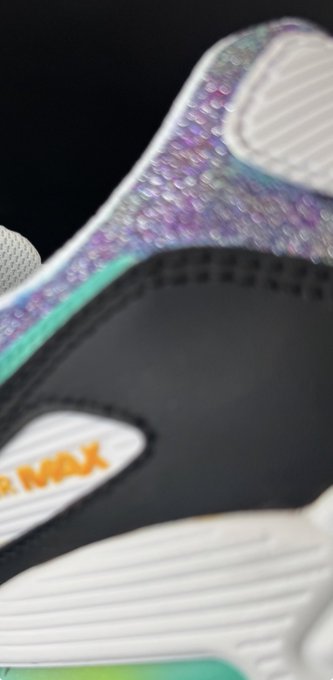 A close-up of a white sneaker with iridescent purple and teal detailing, featuring a black strap with orange text partially visible as 'MIX'.