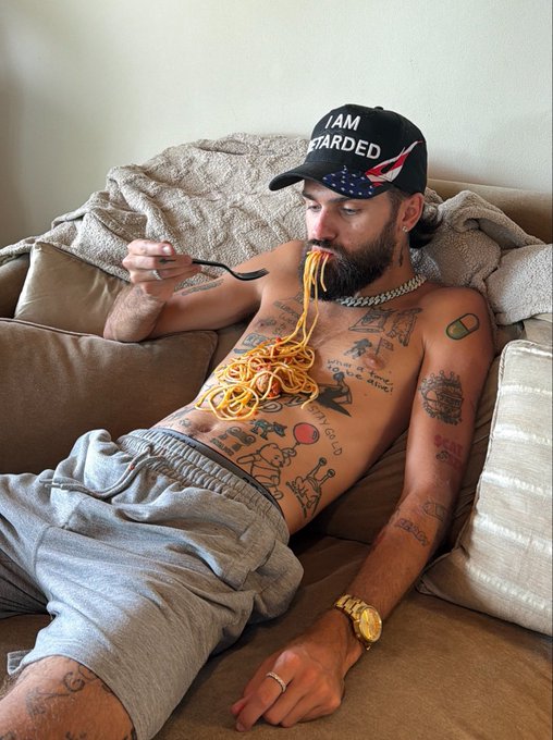 A shirtless, tattooed bearded man wearing an 'I AM RETARDED' cap sits on a beige couch, casually eating a forkful of spaghetti with a gold watch on his wrist.