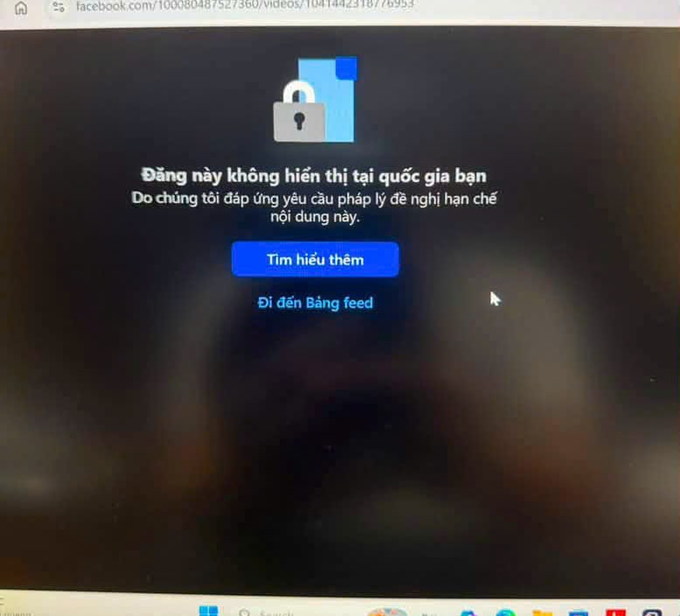 A computer screen displays a Facebook error message in Vietnamese stating 'Đăng này không hiển thị tại quốc gia bạn' with a blue 'Tìm hiểu thêm' button.