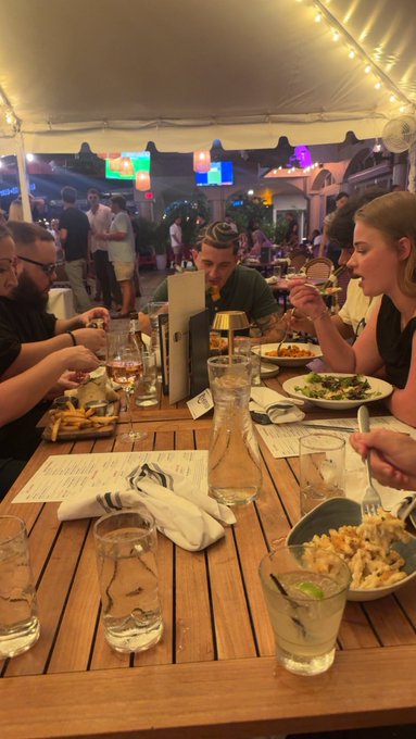 A group of people enjoy dinner together under string lights at an outdoor restaurant, with pasta dishes and drinks scattered across the wooden table, embodying the "family" sentiment referenced in the post. One person sports a flat cap, adding to the casual, friendly atmosphere of the gathering.