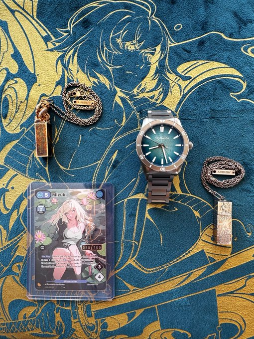 An anime-style merchandise collection on a blue and gold patterned cloth, including a green-dial watch, two pendants on chains, and a holographic 'Mizuki' trading card.