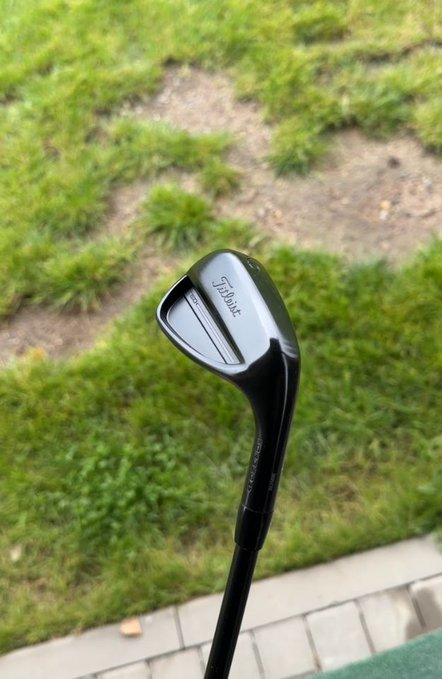 A close-up shows a black golf-club head with “Titleist” printed on it, held above a grassy area and paved edge.