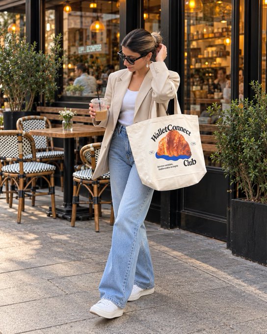 A photograph of a stylish woman walking on a sidewalk outside a café, holding an iced coffee and wearing a tote bag labeled 'WalletConnect Club' featuring a volcano graphic.