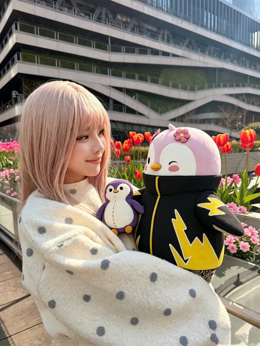 The image features a woman with blonde hair wearing a white sweater, holding a stuffed penguin toy. She has pink flowers in her hair and is smiling at the camera. The background includes a building and some flowers, creating an urban setting for this scene. The caption of the image reads "Me and my Lil Pudgy @LilPudgys".