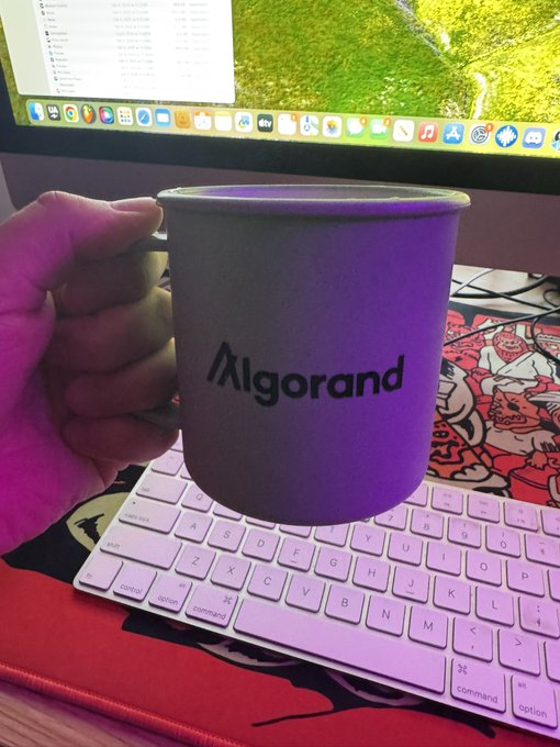 A hand holds a light-colored enamel mug featuring the 'Algorand' logo in front of a computer monitor displaying code and a pink wireless keyboard on a red desk mat.