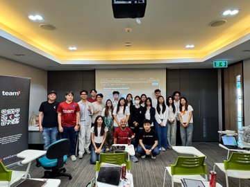 A group of about 17 people posing for a photo in a brightly lit conference or meeting room with a 'teamv' banner on the left.