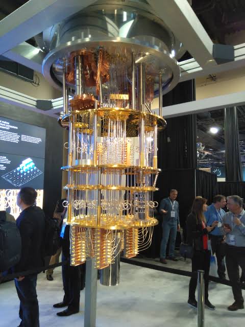 A large gold-colored quantum computer is displayed at a tech conference, with attendees gathered around to view the cylindrical apparatus featuring multiple tiers of intricate circuitry. The accompanying post explains that sufficiently powerful quantum computers could one day threaten cryptographic signature systems, potentially cracking private keys used to secure digital assets like cryptocurrencies.