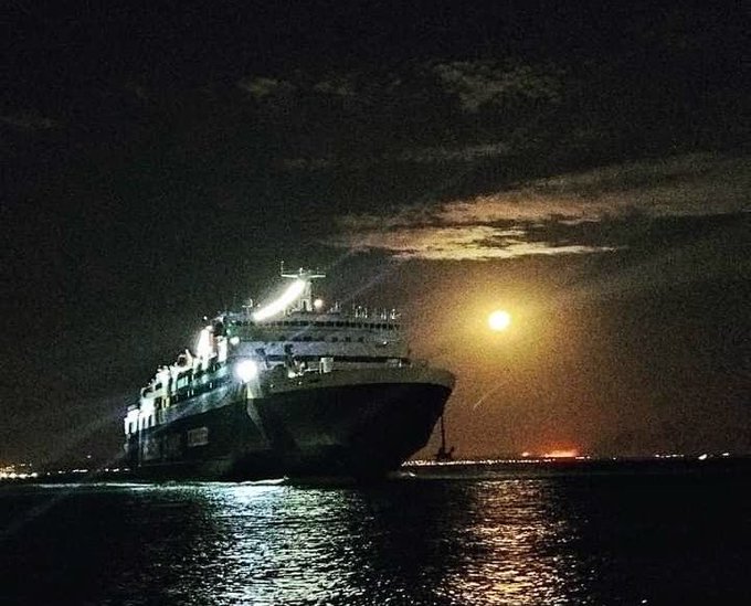 A large illuminated ferry or cruise ship sails through dark waters at night, with a glowing moon casting light on the clouds and reflecting on the sea, while distant city lights twinkle on the horizon.

The image visually reinforces the post's message about the Ethra Ship ($SHIP) project's real-world maritime exposure, pairing the vessel on the water with text describing how the project bridges blockchain staking/governance with RWA-backed ship investments.