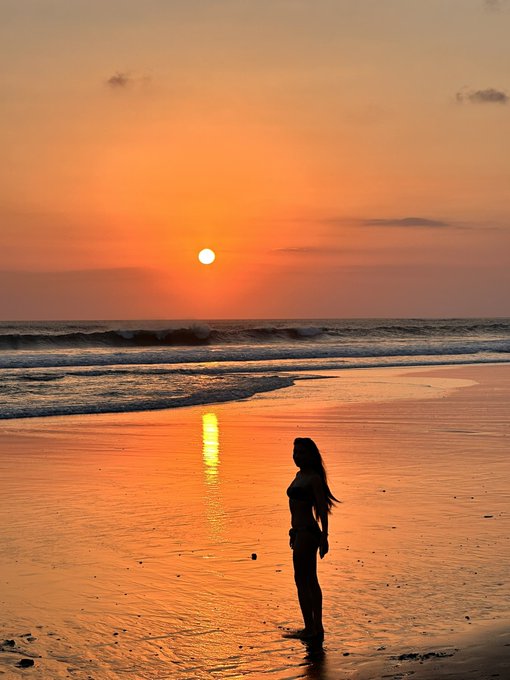 A silhouette of a woman standing on a beach at sunset, with orange and golden hues reflecting on the water.