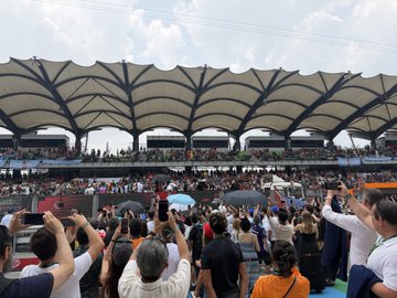 A large crowd gathers under a distinctive umbrella-style canopy at what appears to be the Sepang International Circuit, with spectators holding up phones to capture the action—likely related to the McLaren Formula 1 team sponsored by OKX.
